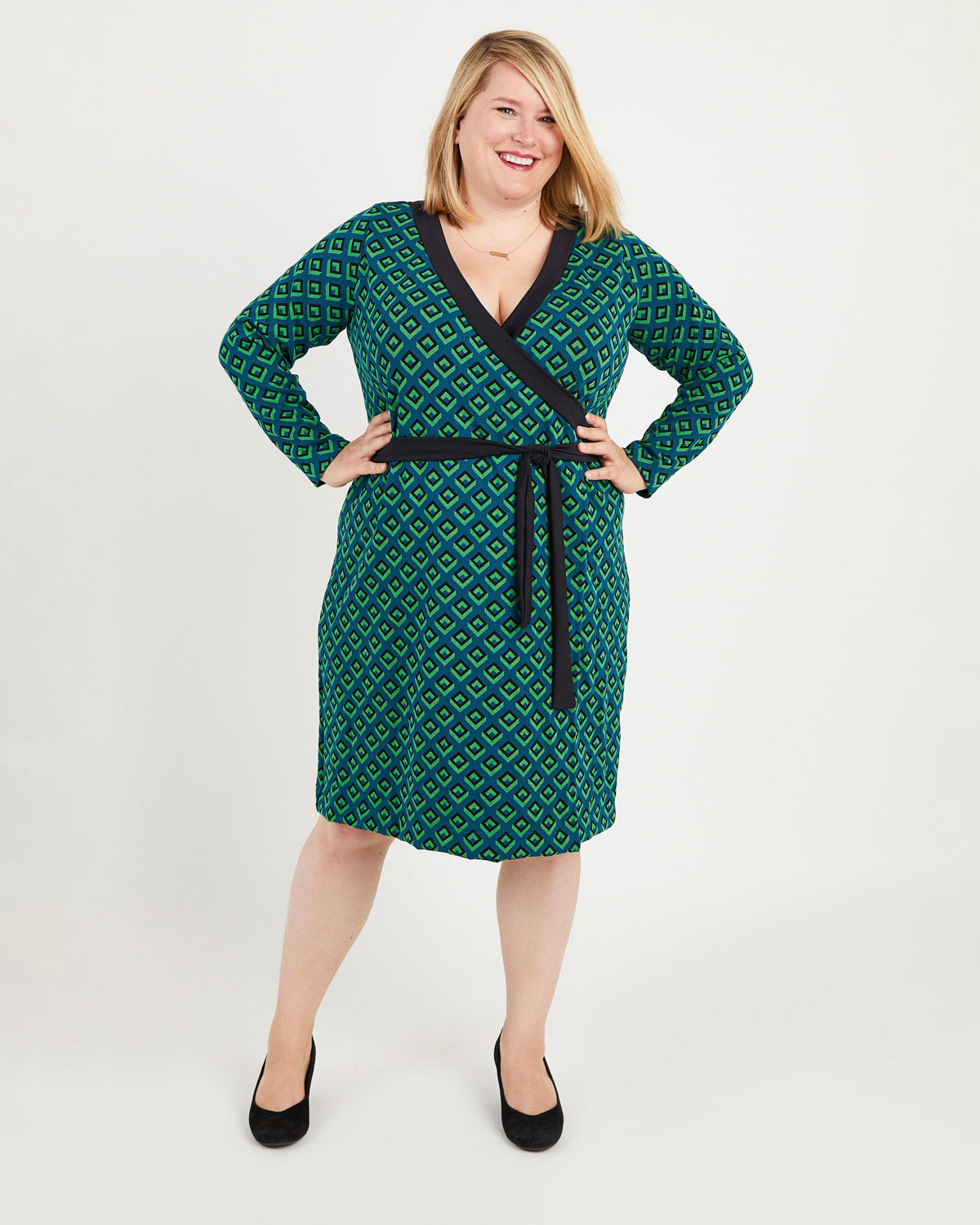 Woman wearing a green patterned dress with a belt on a white background. This dress is the Appleton Wrap Dress sewing pattern from Cashmerette.