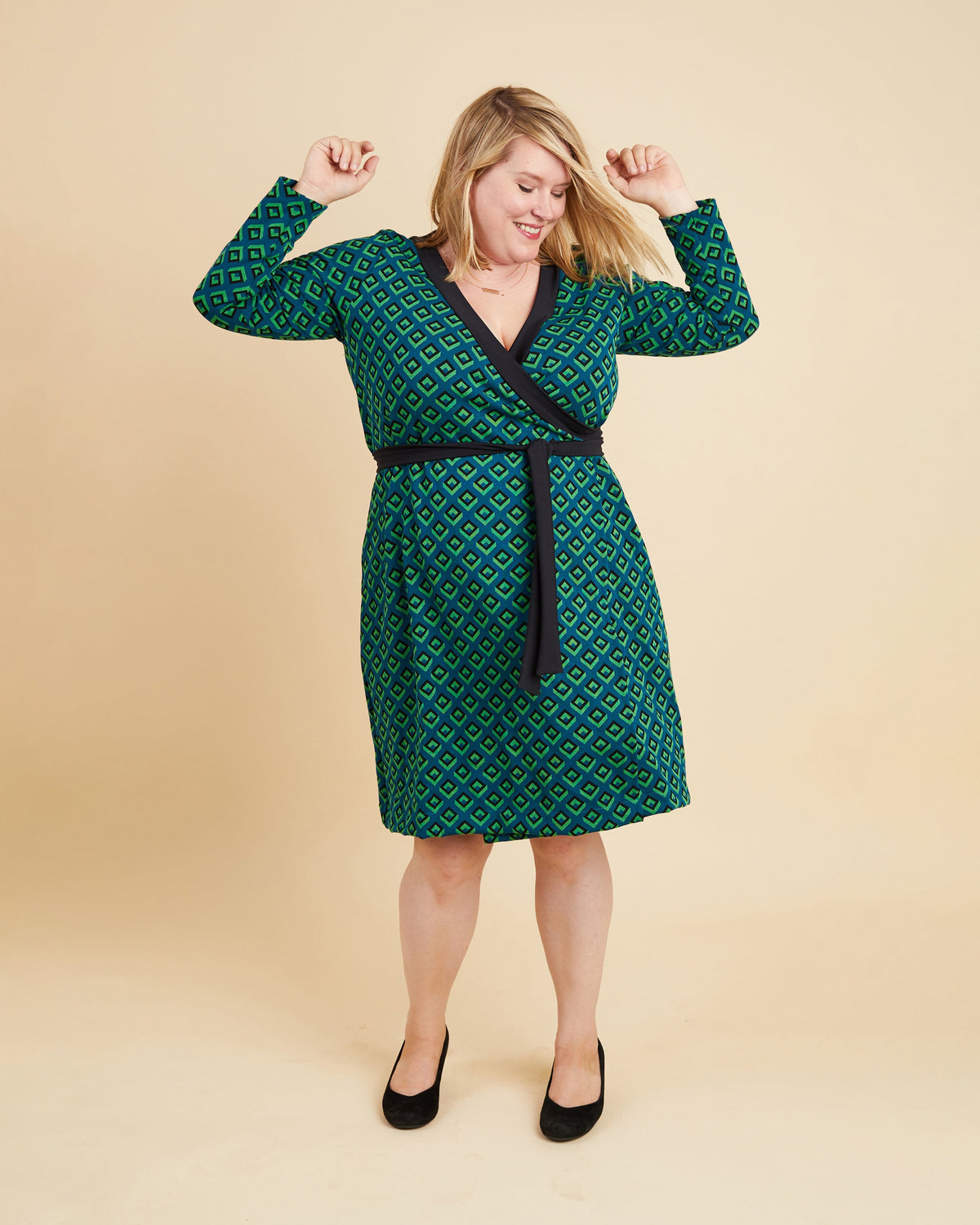 Woman wearing a green patterned dress with a belt on a beige background. This dress is the Appleton Wrap Dress sewing pattern from Cashmerette.