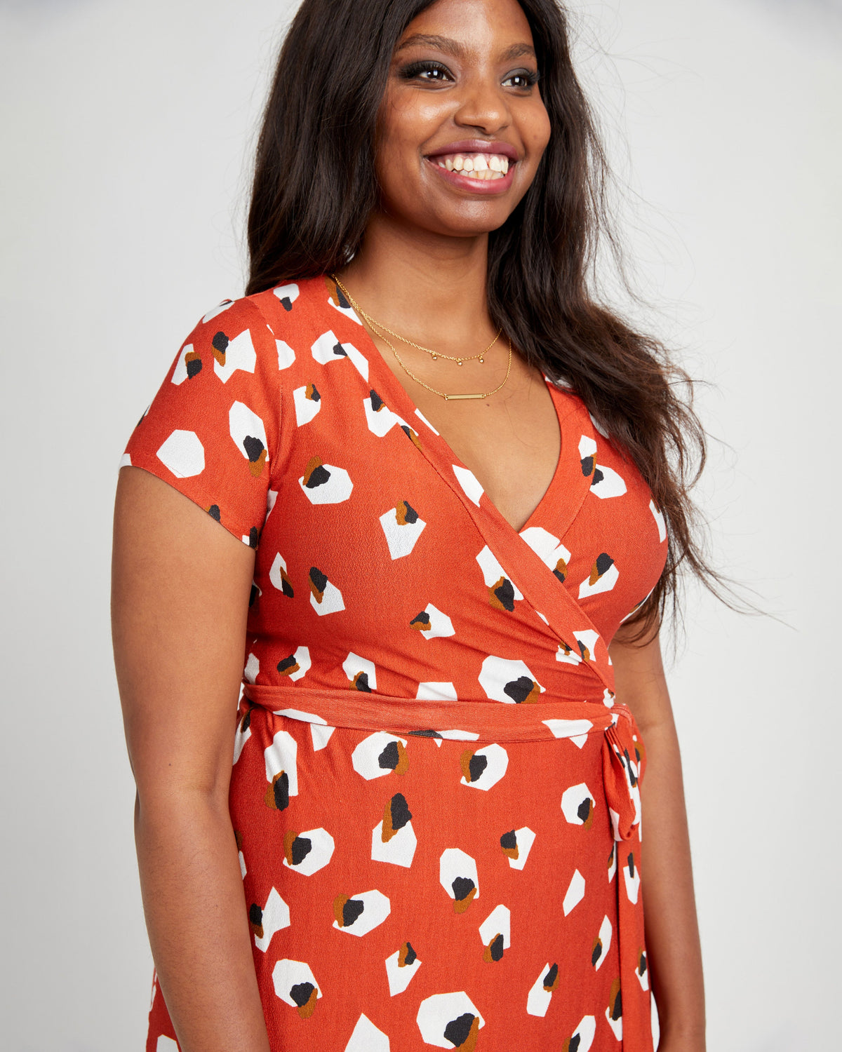 Woman wearing an orange dress with a pattern of white and black shapes on a plain background. This dress is the Appleton Wrap Dress sewing pattern from Cashmerette.