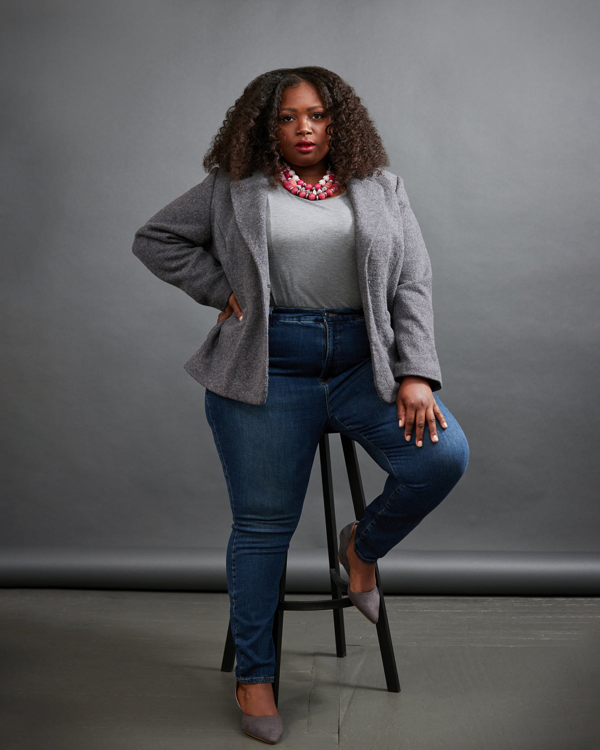 Woman sitting on a stool wearing a gray cardigan, white shirt, blue jeans, and gray shoes against a gray background. This blazer is the Auburn Blazer sewing pattern from Cashmerette.