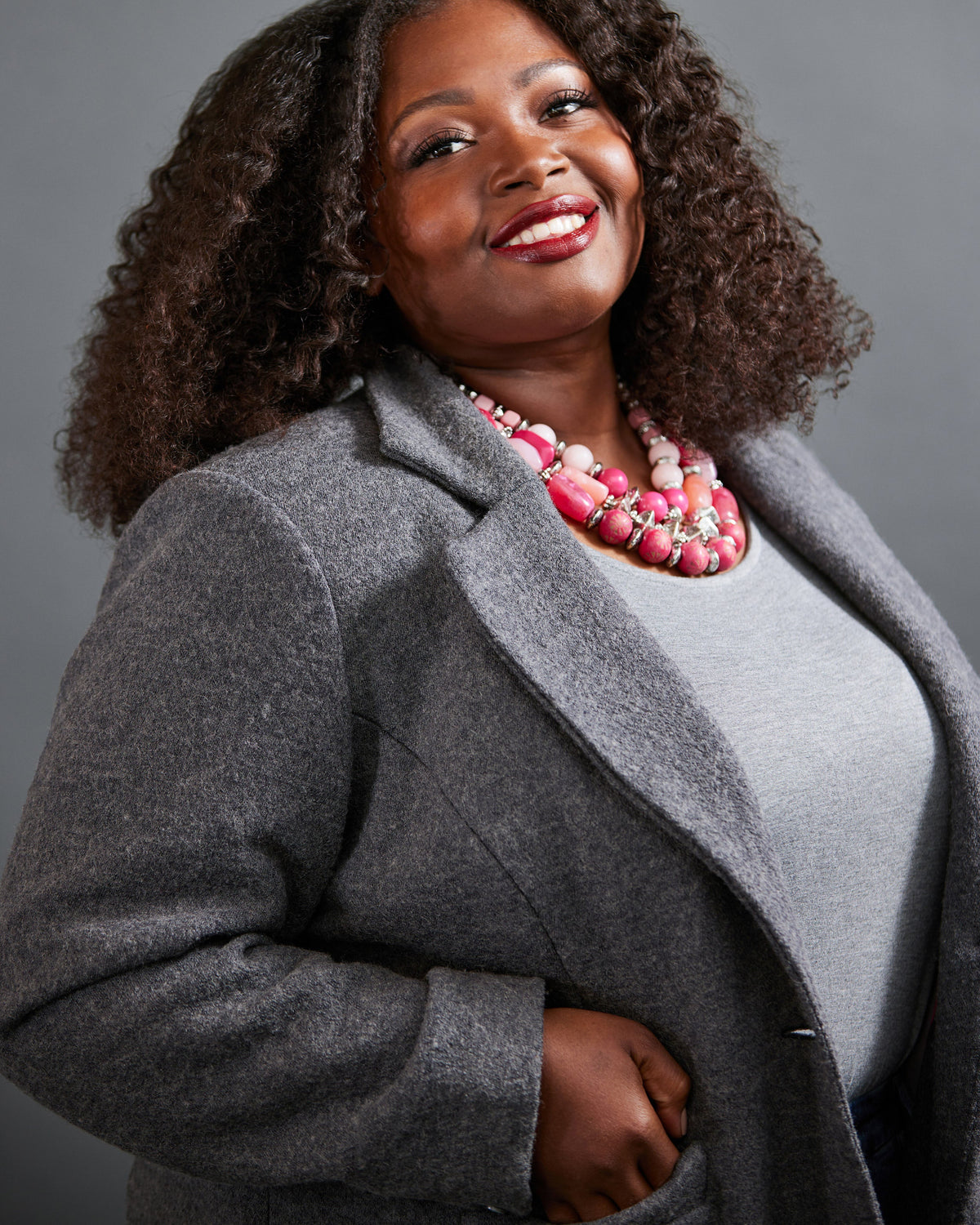 Woman wearing a gray coat and pink necklace against a gray background. This blazer is the Auburn Blazer sewing pattern from Cashmerette.