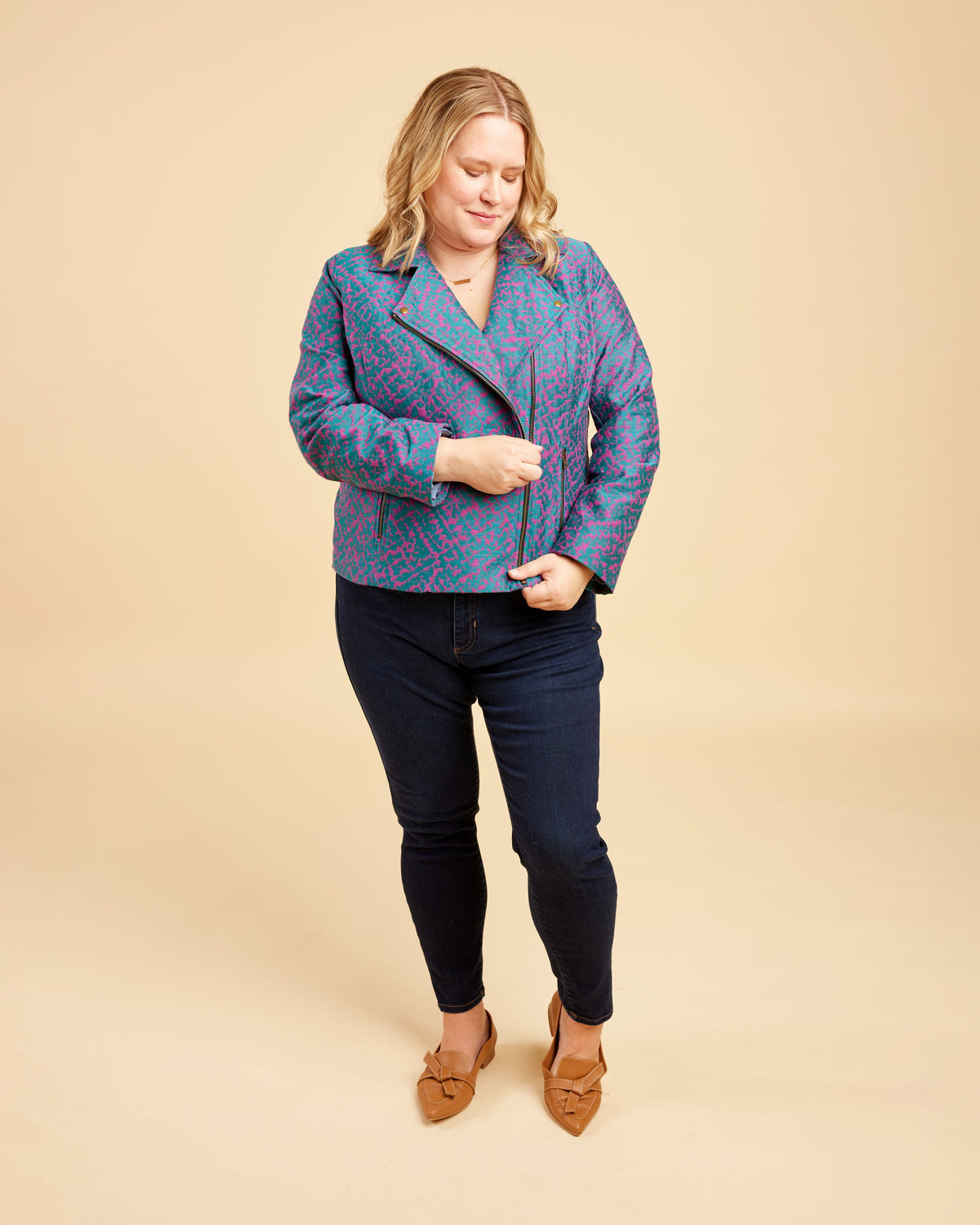 Woman wearing a blue patterned jacket and dark jeans on a beige background. This biker jacket is the Canton Moto Jacket sewing pattern from Cashmerette.