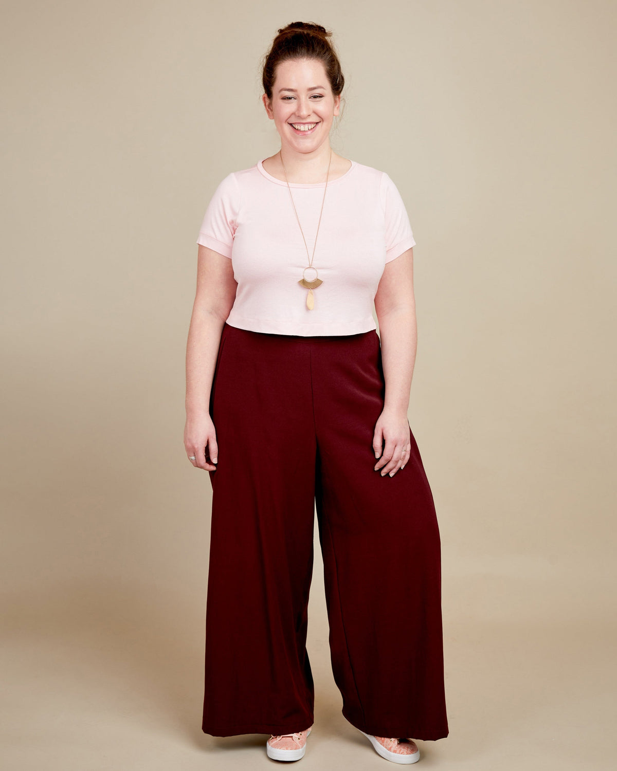 Woman wearing a pink top and burgundy wide-leg pants on a beige background. This t-shirt is the Concord T-Shirt sewing pattern from Cashmerette.