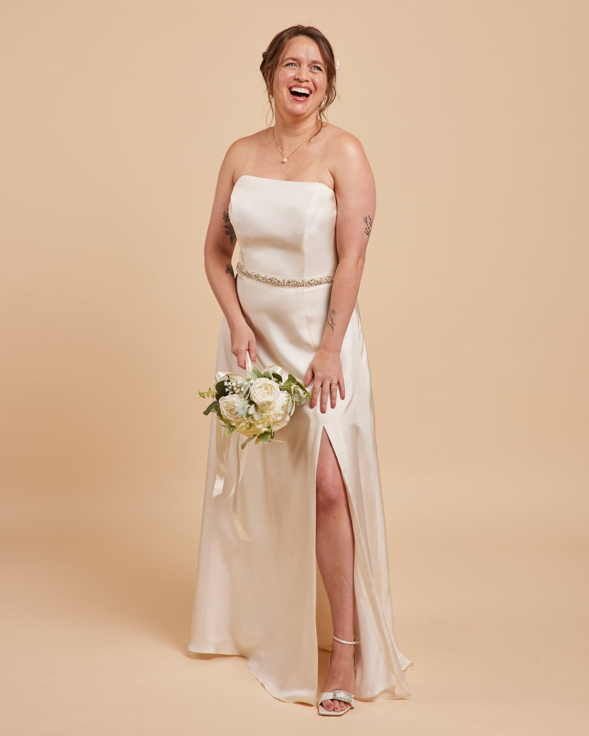 Woman in a strapless white wedding dress with a high slit, holding a bouquet on a beige background. This dress is the Ellington Strapless Dress sewing pattern from Cashmerette.