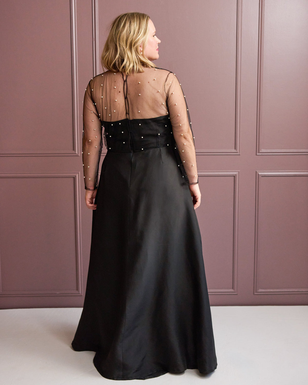 Woman wearing a black evening gown with sheer sleeves against a pink paneled wall. This is the Ellington Dress sewing pattern from Cashmerette.