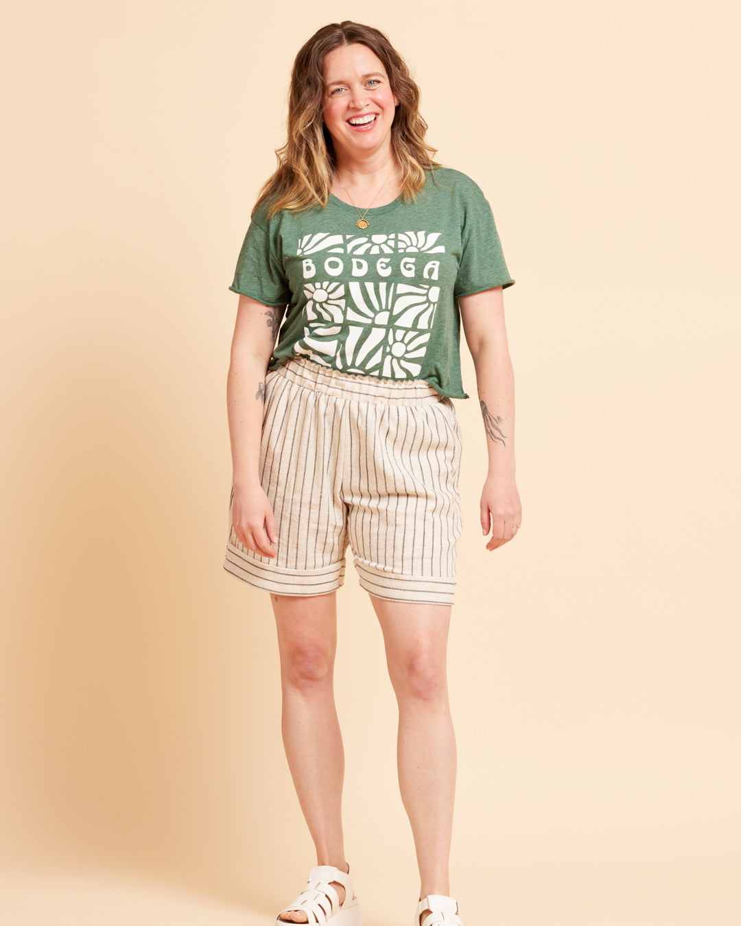 Woman wearing a green t-shirt with 'Bodega' print and beige shorts on a beige background. These shorts are the Everett Pants & Shorts sewing pattern from Cashmerette.