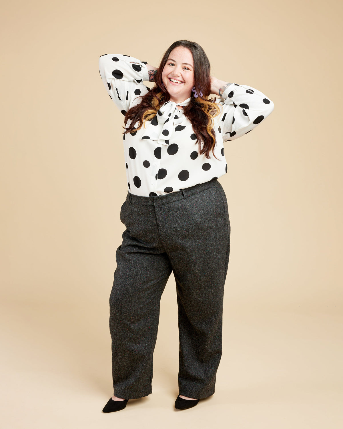 Person wearing a polka dot blouse and gray pants on a beige background. These pants are the Meriam Trousers sewing pattern from Cashmerette.