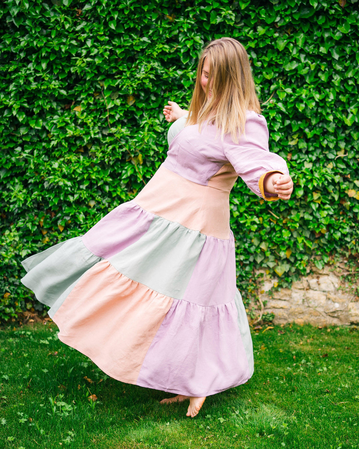 Woman in a pastel-colored dress twirling in front of a green hedge.. This dress is the Roseclair Wrap Dress sewing pattern from Cashmerette.