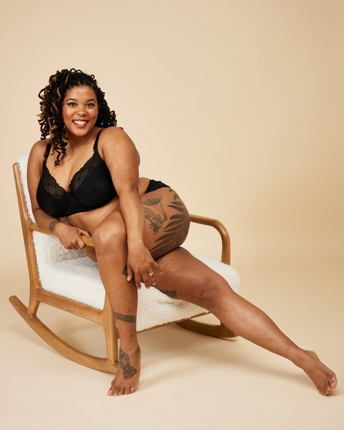 Woman sitting on a rocking chair wearing black lingerie against a beige background. This bra is the Willowdale Bra sewing pattern from Cashmerette.
