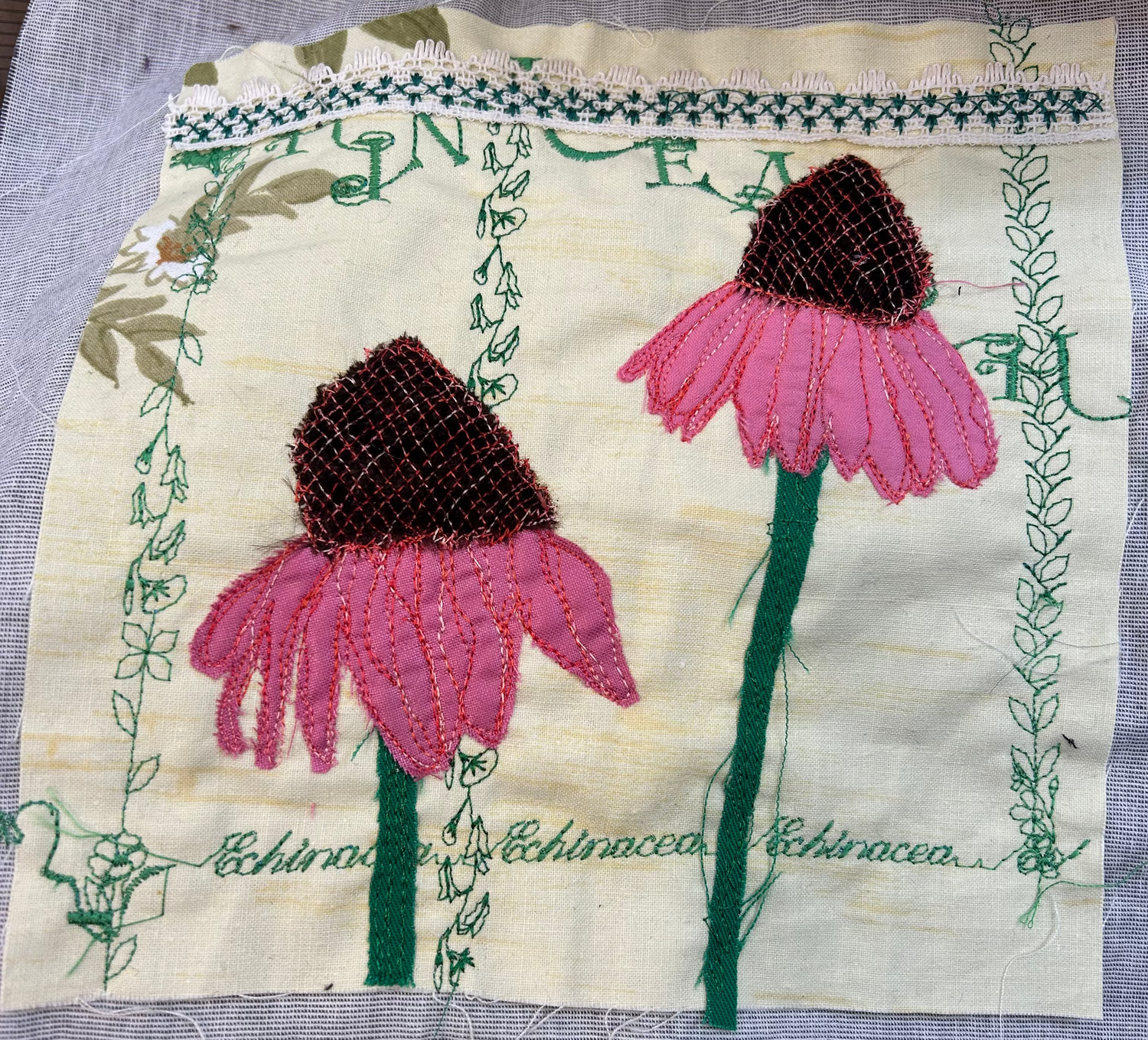 A square embroidered fabric patch featuring two pink echinacea flowers with dark brown centers, green stems, and green decorative borders and script text on a cream background, placed on a gray textile surface.