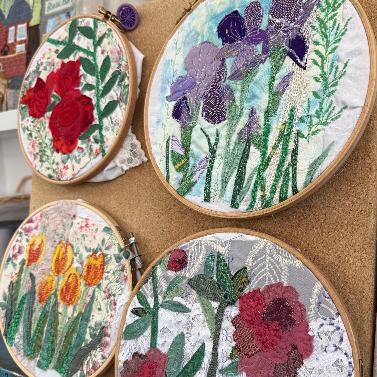 Four circular embroidery hoops displaying colorful floral designs, including red poppies, purple irises, yellow tulips, and red roses, arranged on a cork board in a craft workspace.