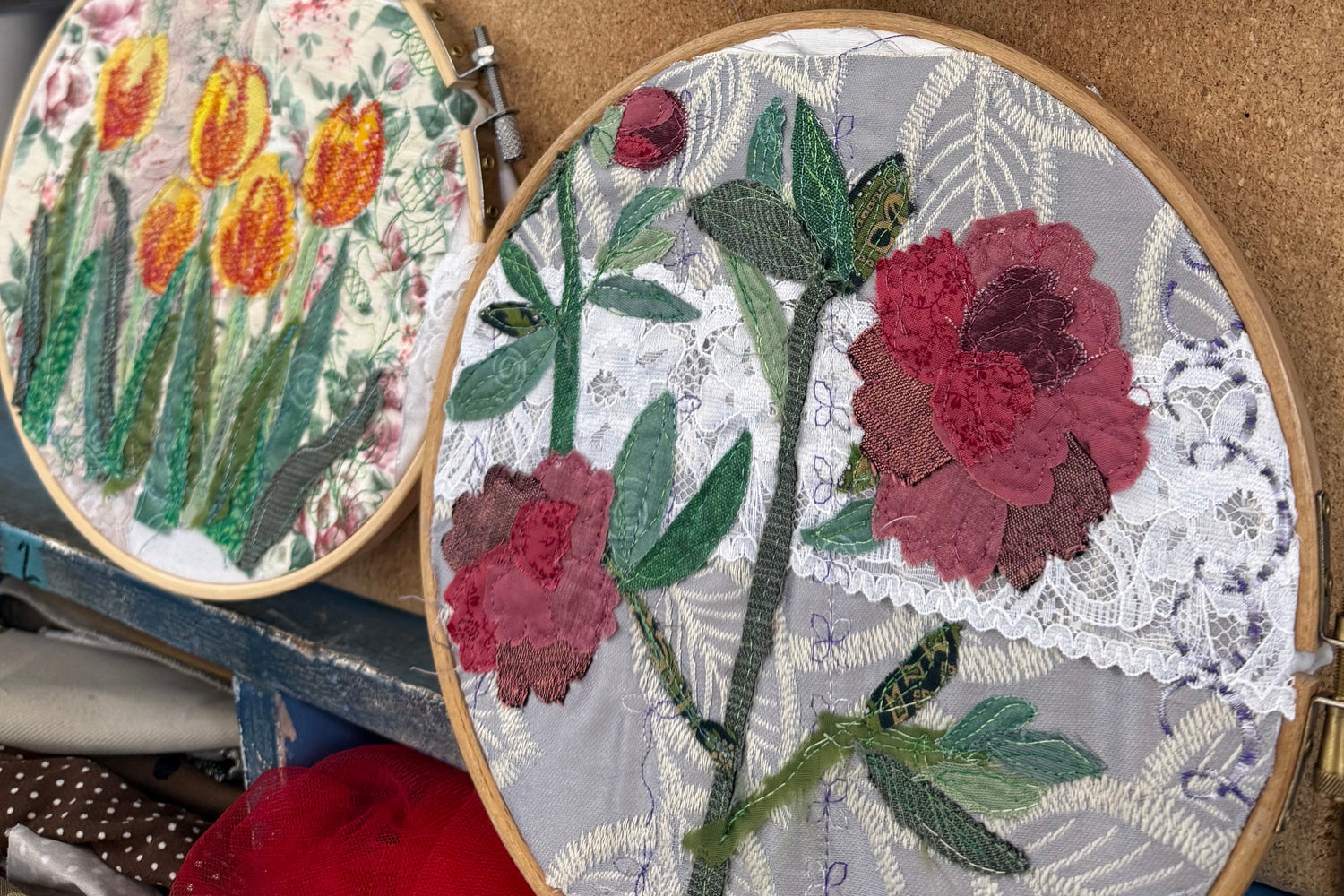 Four circular embroidery hoops displaying colorful floral designs, including red poppies, purple irises, yellow tulips, and red roses, arranged on a cork board in a craft workspace.