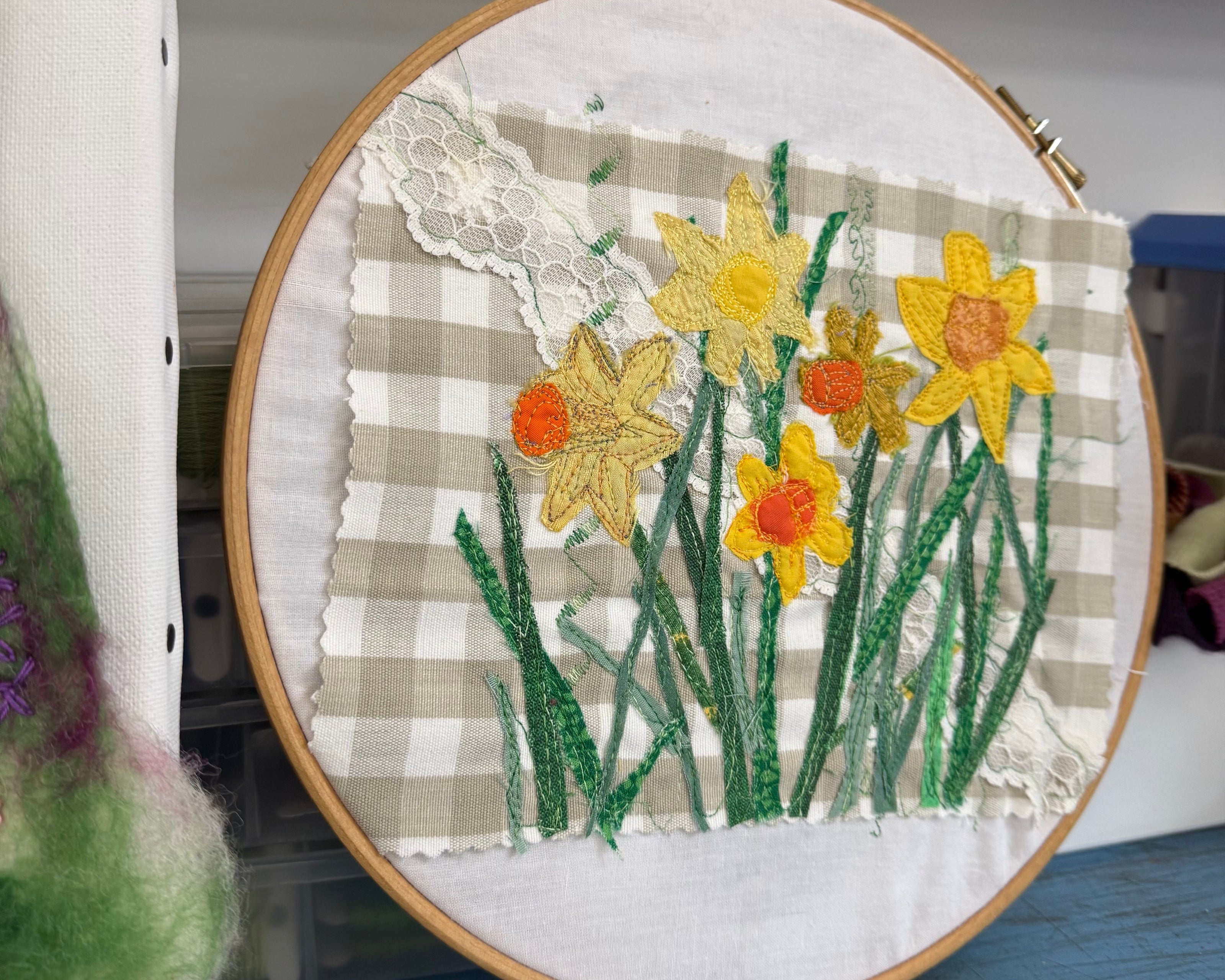 A round embroidery hoop holding a handmade textile artwork with embroidered yellow daffodils, green stems, a beige-and-white gingham background, and a white lace applique, displayed on a blue wooden surface.