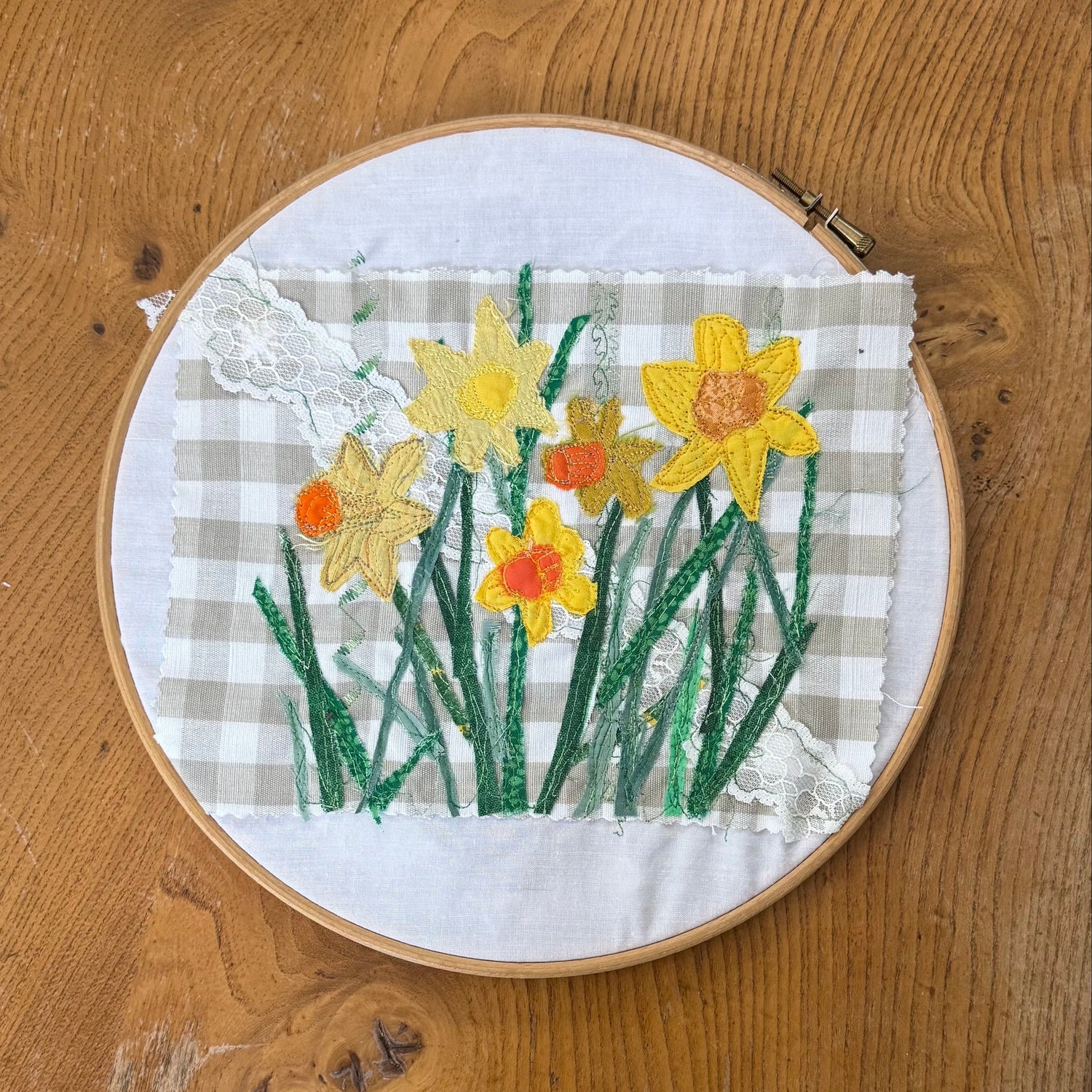A circular embroidery hoop containing a floral patchwork design with yellow and orange daffodils on a beige-and-white gingham background, placed on a wooden tabletop.