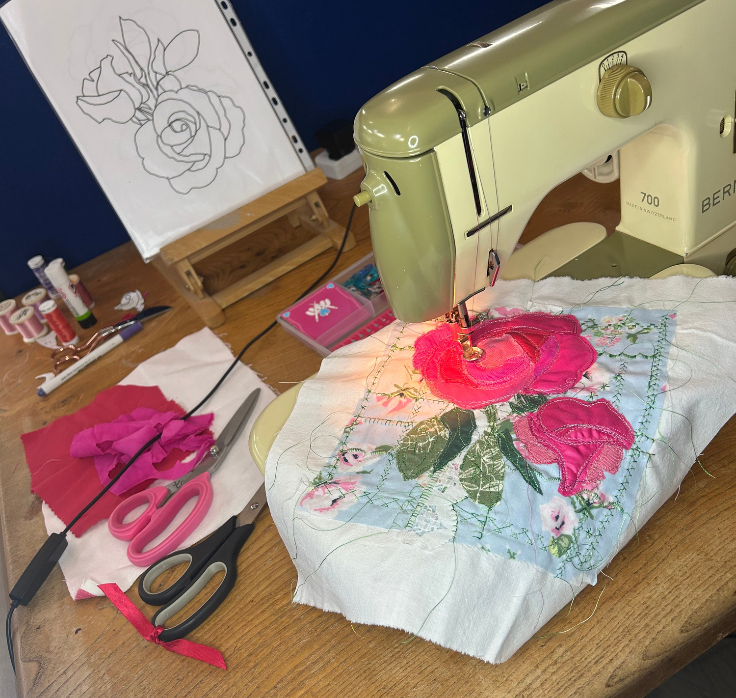 A vintage cream-colored sewing machine on a wooden table next to a piece of floral fabric, with scissors, thread, and an embroidery frame visible in a craft workspace.