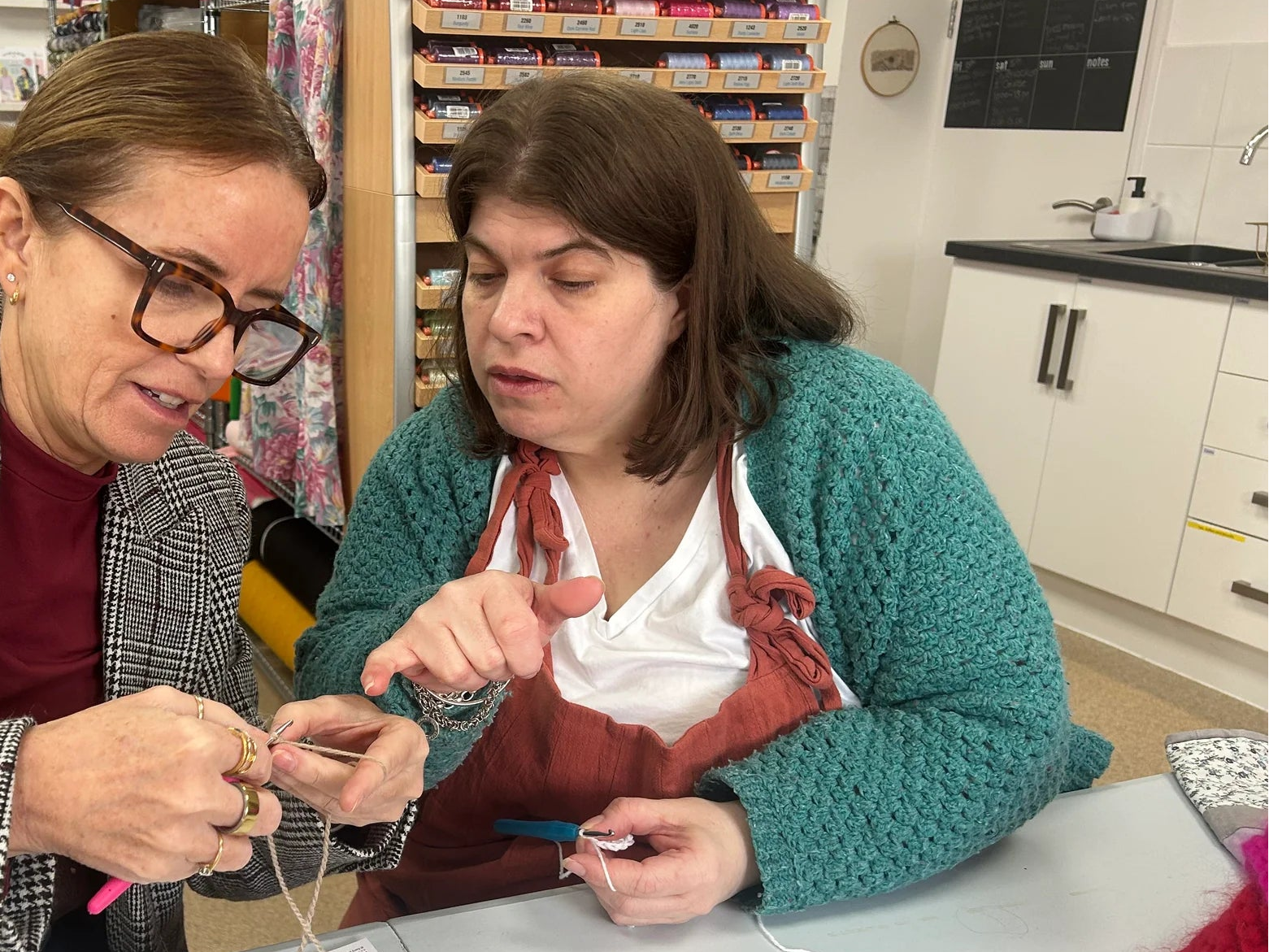 Two women seated at a table in a craft or sewing supply shop, working together on a knitting or crocheting project with yarn and a pattern sheet visible.