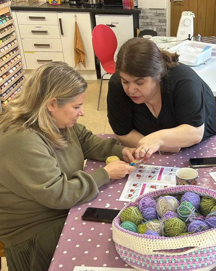 Four women gathered around a table in a craft room, working on crochet or knitting projects. A woven basket filled with colorful balls of yarn sits on a pink polka-dot tablecloth, surrounded by pattern sheets, cups, and crafting supplies.