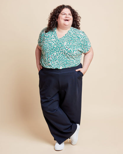 Woman wearing a floral top and dark pants on a beige background. This is the Grafton Dress, Top & Skirt sewing pattern from Cashmerette.