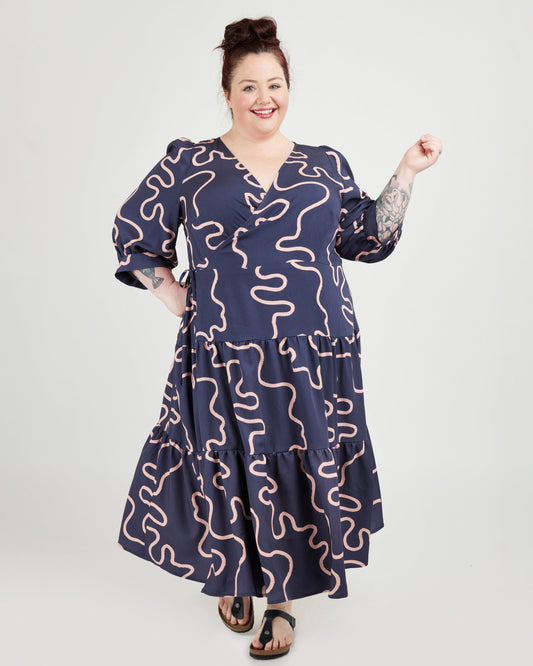 Woman wearing a navy dress with pink wavy patterns on a white background. This dress is the Roseclair Wrap Dress sewing pattern from Cashmerette.